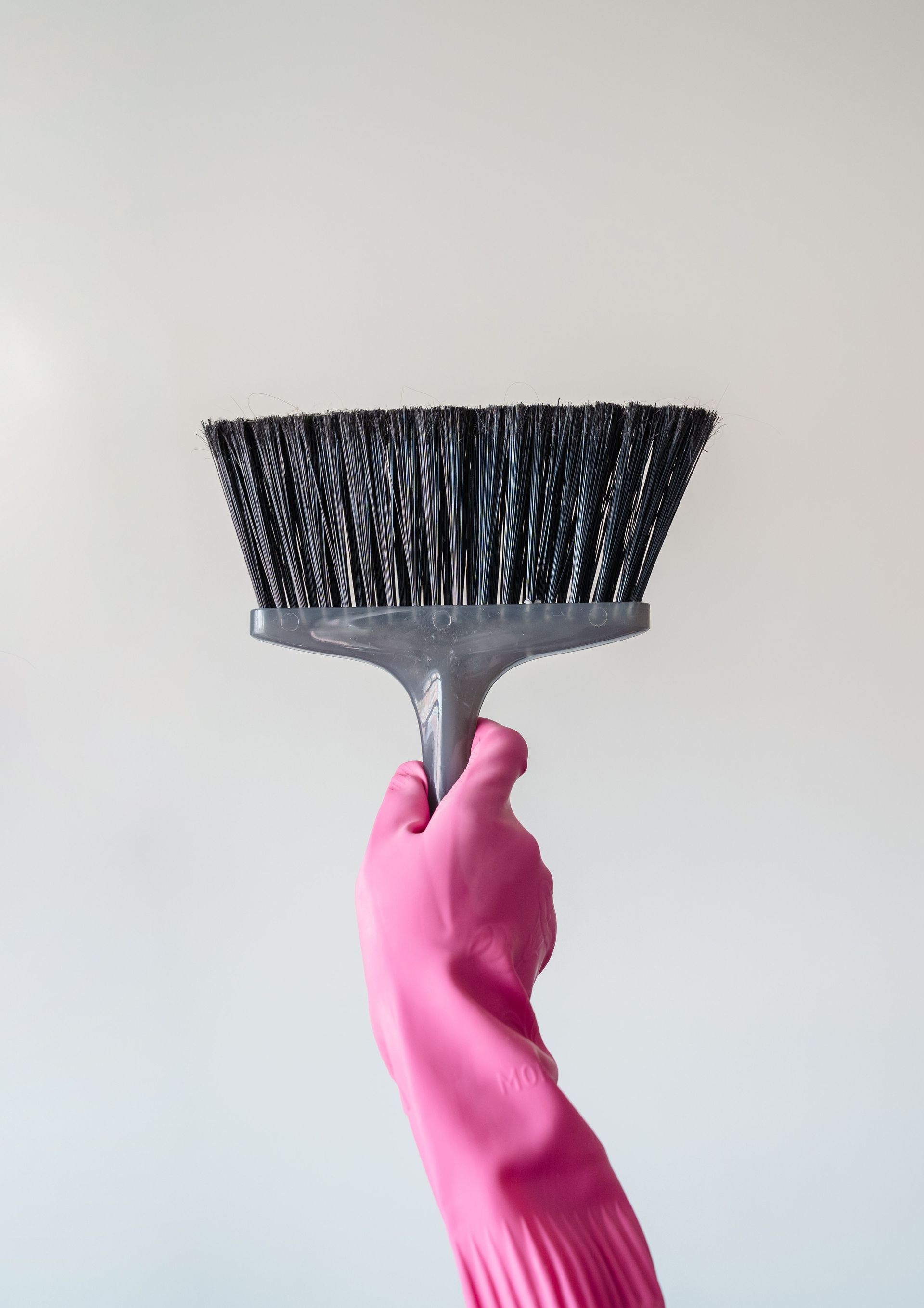 A close-up of a person holding a broom wearing a pink cleaning glove against a plain background.
